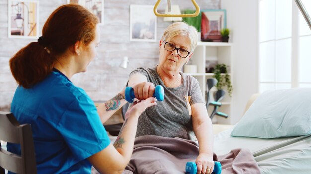 Revealing shot of female nurse helping an elderly woman to recover her muscles after an accident. She is lying in a hospital bed in a retirement home