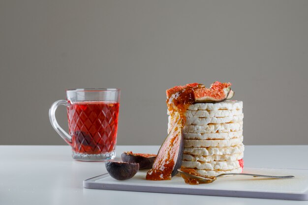 Rice cakes with figs, jam, tea, teaspoon on cutting board on white and grey,