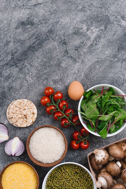 Rice grains; mung beans; puffed rice cake; polenta; cherry tomatoes; egg; mushroom and halved onion against concrete backdrop