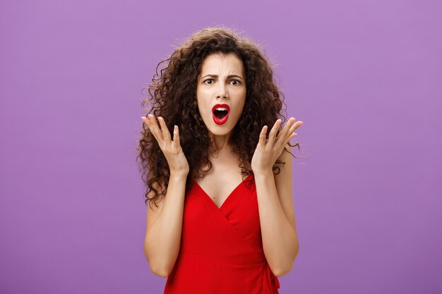 Rich arrogant and snobbish european woman with curly hairstyle in red evening dress arguing with made frowning looking confused and displeased shaking palms in disappointment posing over purple wall.