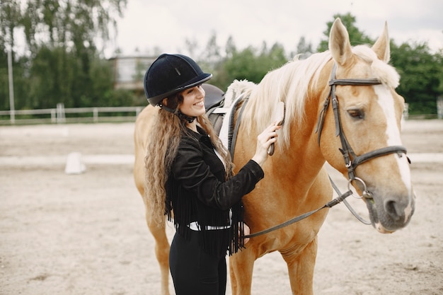 ライダーの女性は牧場で彼女の馬をとかします。女性は長い髪と黒い服を着ています。彼女の茶色の馬に触れる女性の乗馬。