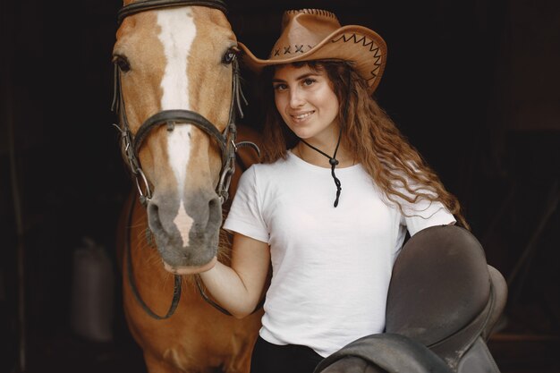 Rider woman holding a saddle in a stable. Woman has long hair and white t-shirt