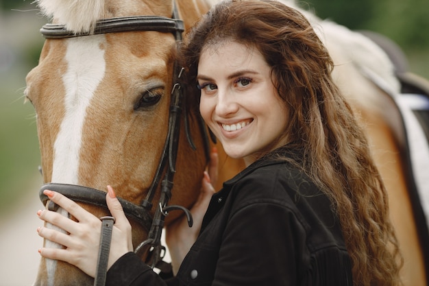 Женщина всадника смотря камеру. У женщины длинные волосы и черная одежда. Женский наездник трогает поводья лошади.