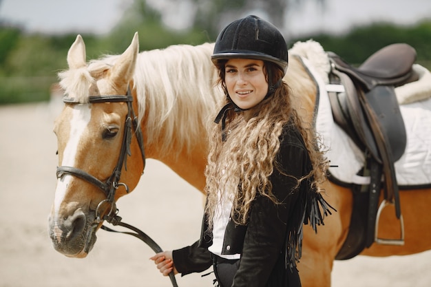 Donna del cavaliere che esamina la macchina fotografica. la donna ha i capelli lunghi e vestiti neri. equestre femminile che tocca le redini di un cavallo.