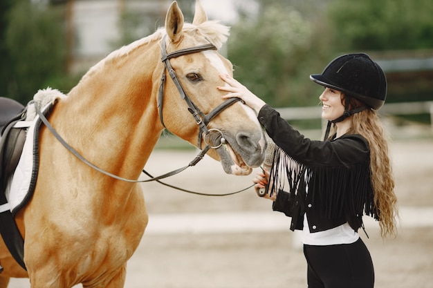 Rider woman talking to her horse on a ranch. Woman has long hair and black clothes. Female equestrian touching her brown horse.
