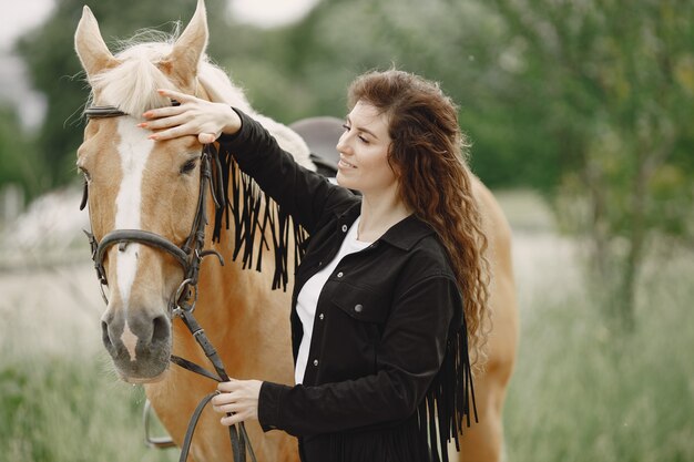 Женщина-всадник разговаривает со своей лошадью на ранчо. У женщины длинные волосы и черная одежда. Женский наездник трогает поводья лошади.