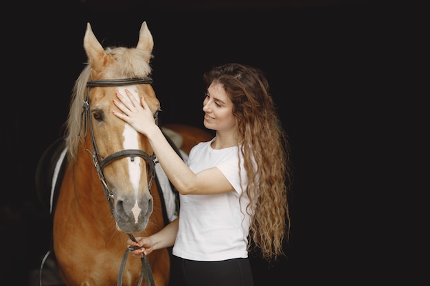 Free photo rider woman talking to her horse in a stable. woman has long hair and white t-shirt. background is dark and black.