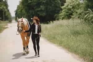 Foto gratuita donna del cavaliere che cammina con il suo cavallo su una strada. la donna ha i capelli lunghi e vestiti neri. equestre femminile che tiene le redini di un cavallo.