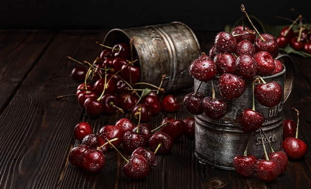 Ripe cherries in drops of water, is in vintage metal mugs. Dark wood background, selective focus. Fresh harvest of juicy cherries