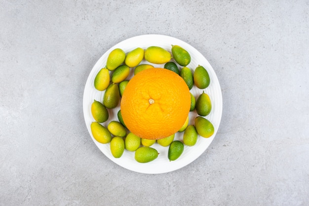 Спелый апельсин с кучей кумкватов на белой тарелке.