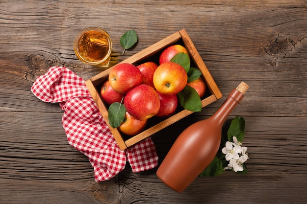 Спелые красные яблоки в деревянной коробке с веткой белых цветов, стаканом и бутылкой сидра на деревянном столе. вид сверху с пространством для вашего текста.