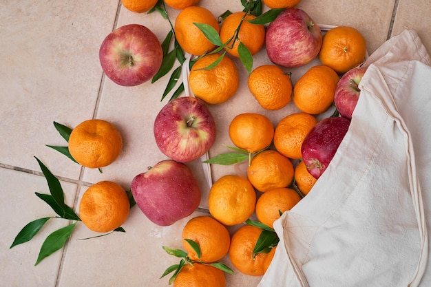 Free photo ripe tangerines and red apples in a reusable eco bag on a ceramic floor fruit delivery healthy food vitamins top view of juicy fruits