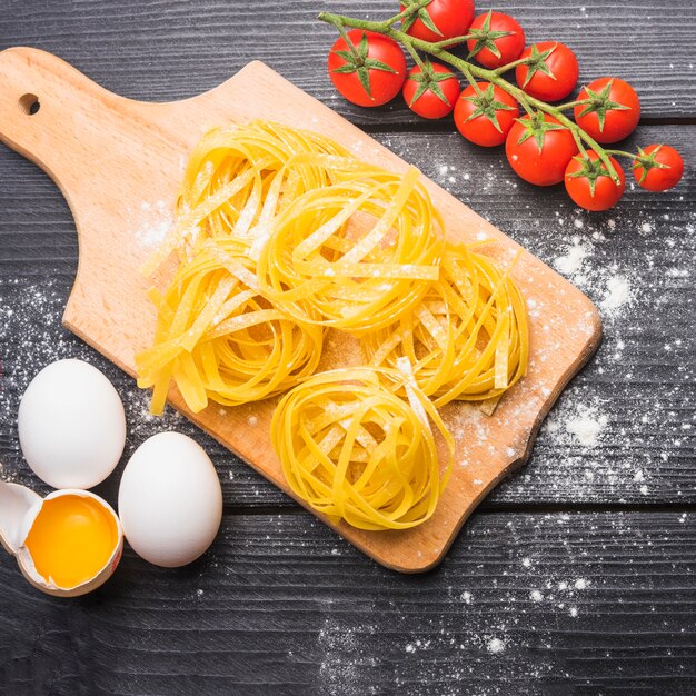 Ripe tomatoes; raw tagliatelle with broken and whole eggs on wooden plank