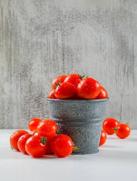 Ripe wet tomatoes in a mini backet on a grungy wall and white surface. side view.