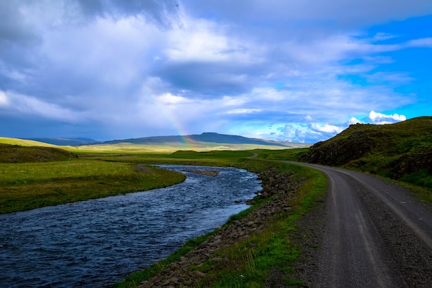River in the middle of a road and grassy field with a rainbow in the distance at daytime