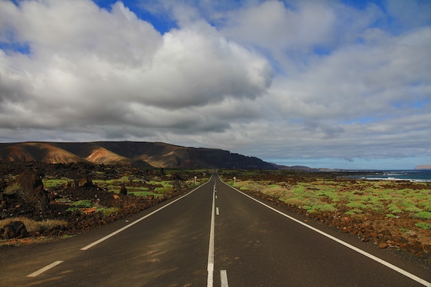 Road in the middle of a grassy field with a mountain in the distance under a cloudy sky