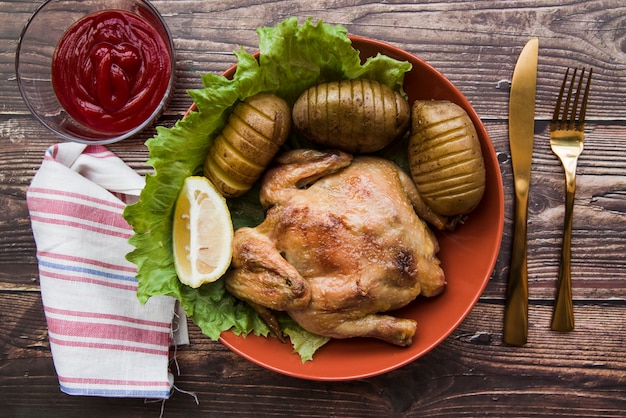 Roasted chicken in bowl with potato; knife; napkin; lemon; sauce and fork on desk