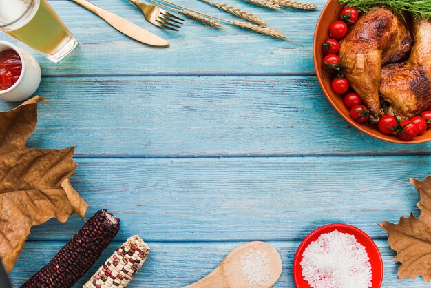 Roasted chicken leg; in bowl with salt; leaves; beer; fork; butterknife; corn on wooden table