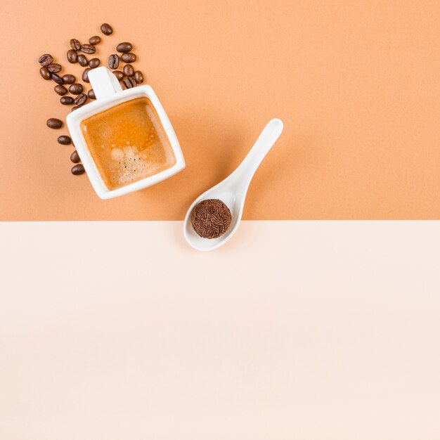 Жареные кофейные бобы; чашка кофе и шоколадный шар в ложке на бежевом двойном фоне