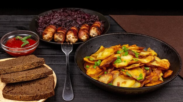 鍋にローストポテト、木製のサービングプレートにライ麦の黒パン、ベーコンに自家製ソーセージ、黒の素朴な背景にキャベツの煮込み。