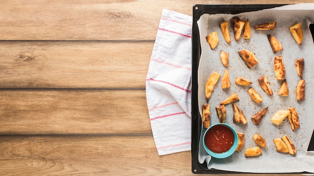 Roasted potato pieces with ketchup in tray over wooden table