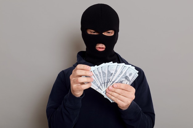 Free photo robber man dressed in black hoodie stands with disguised face and holds a lot of money in his hands