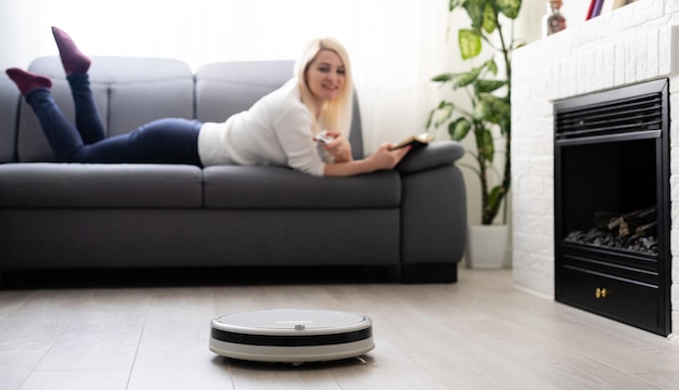 Robotic vacuum cleaner cleaning the room while woman sitting on sofa, closeup.