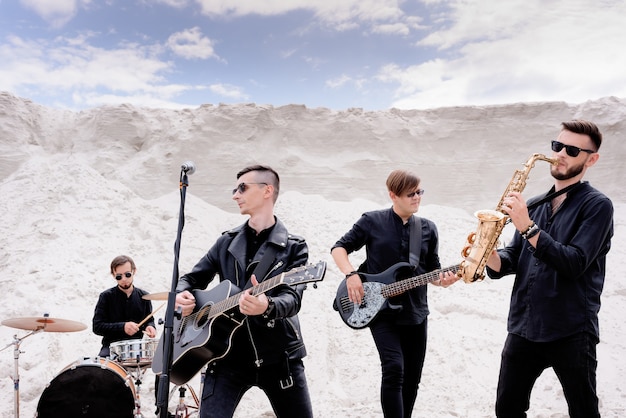 Foto gratuita banda musicale rock che si esibisce in un concerto sulla spiaggia. uomini vestiti con abiti neri in stile rock e occhiali da sole neri