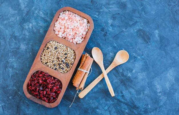 Rock salt in wooden plate with dried barberries, quinoa, black pepper, cinnamon sticks, spoons flat lay on blue grunge background