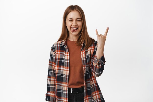 Rock on. Sassy young woman show tongue, wink and heavy metal horns gesture, enjoy party, music event, having fun and celebrate, standing against white background