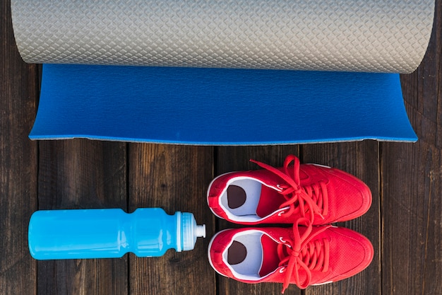 Free photo rolled up exercise mat with water bottle and pair of sport shoes on wooden table