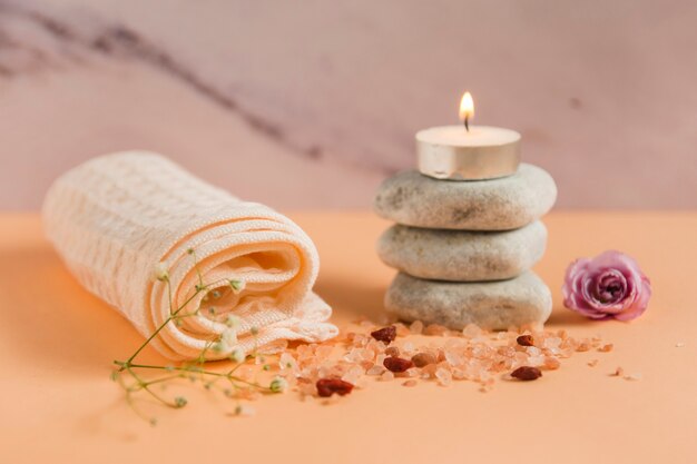 Rolled up towel; lighted candle over the spa stones; rose and himalayan salts on peach colored backdrop