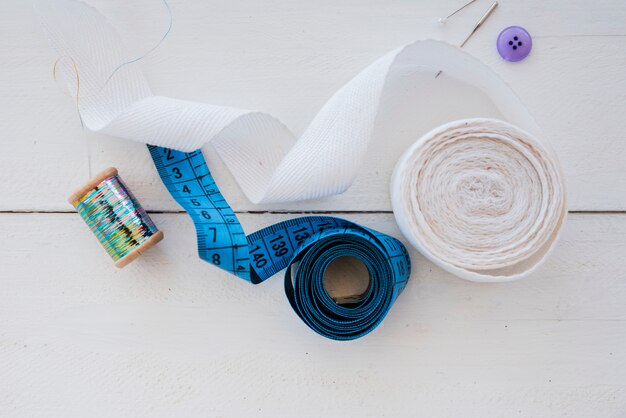 Rolled up white ribbon; measuring tape; button; needle and silver spool on white wooden desk