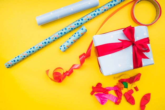 Free photo rolledup gift paper; red ribbon; deflated balloons and present on yellow background