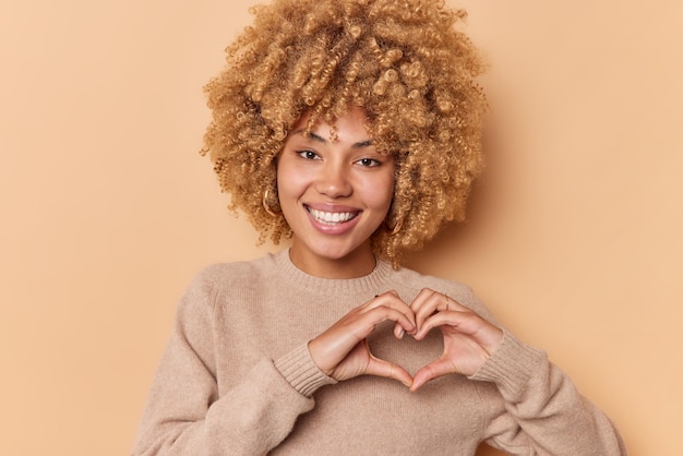 Concetto di romanticismo. la giovane bella donna dai capelli ricci positivi fa il gesto del cuore mostra l'amore sorride teneramente guarda la fotocamera indossa un maglione casual isolato su sfondo beige. sii il mio san valentino.