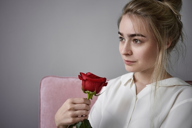 Free photo romance, love and natural beauty concept. close up cropped view of beautiful romantic young female with fair hair wearing white blouse sitting isolated, smelling red rose on valentine's day