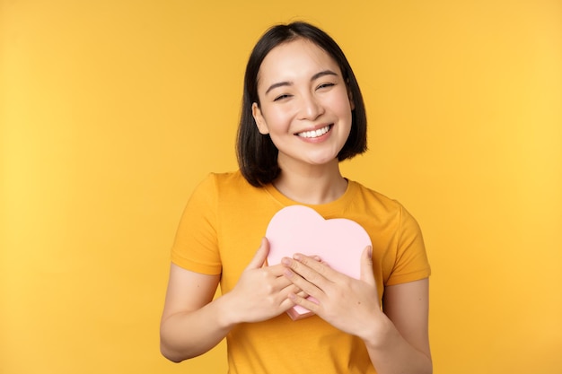 로맨스와 발렌타인 데이 큰 하트 카드를 들고 노란색 배경 위에 서서 웃고 있는 행복한 아름다운 아시아 여성
