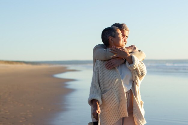 Romanic senior couple spending time at seashore at sunset, enjoying fantastic sea view. Grey-haired man hugging his wife while standing behind. Romance, retirement, holiday concept