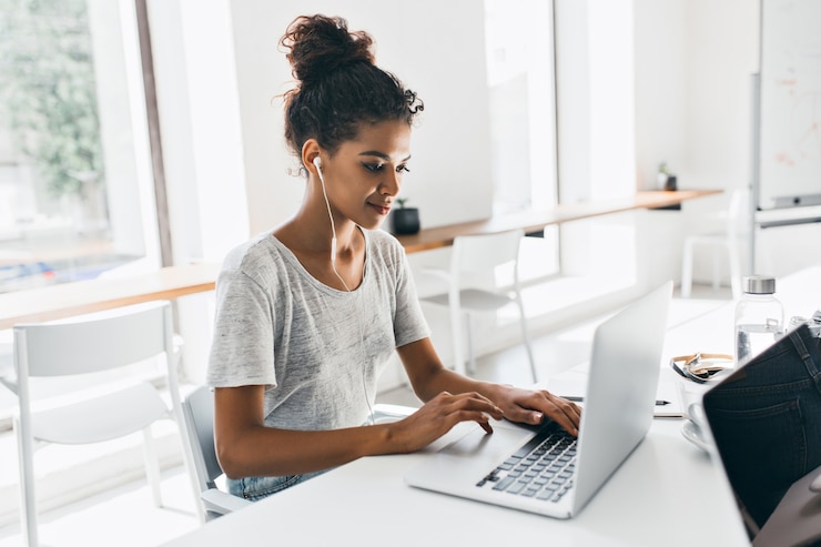 Free Photo romantic african woman with trendy hairstyle sitting at her workplace and analysing data. indoor portrait of black female student working with laptop before exam.