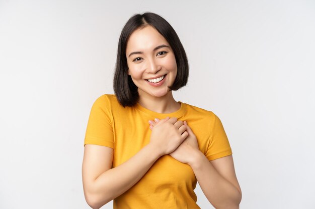 Romantic asian girfriend holding hands on heart smiling with care and tenderness standing in yellow tshirt over white background