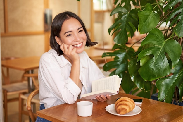 クロワッサンを食べてコーヒーを飲みながらカフェで本を読んで座っているロマンチックなアジアの女性