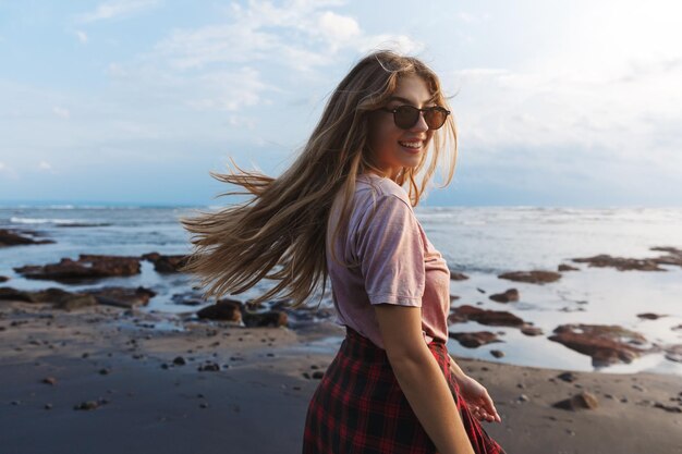 Foto gratuita viaggiatore zaino in spalla romantico attraente hipster ragazza viaggiatore godendo le vacanze estive girarsi dietro per guardare indietro con sorriso camminando lungo la spiaggia vulcanica di sabbia nera al resort tropicale dell'indonesia