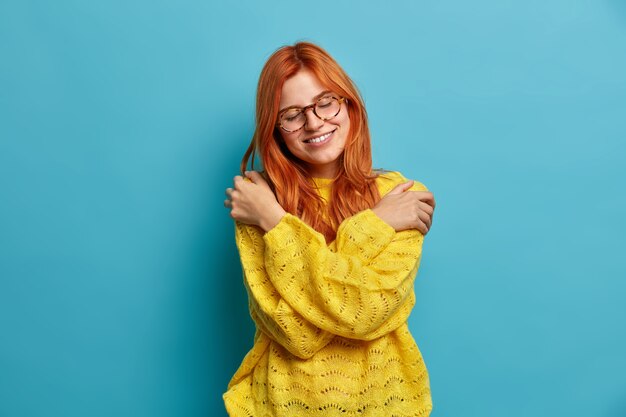 Romantic beautiful redhead woman embraces herself expresses self love and care tilts head smiles gently closes eyes hugs own body wears warm yellow sweater expresses calmness.