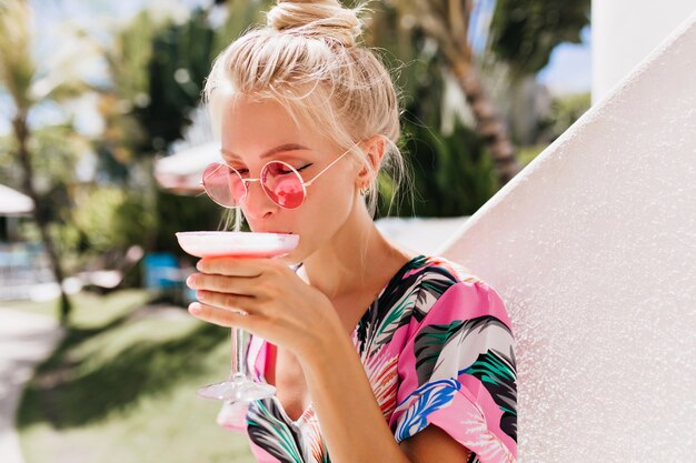 Romantic blonde woman wears sunglasses drinking cocktail with pleasure.