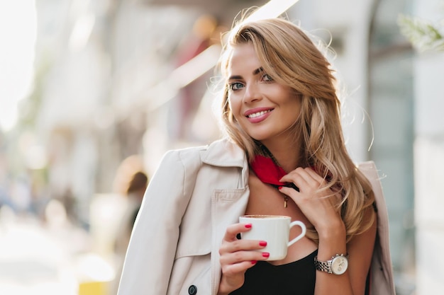 Romantica ragazza dagli occhi azzurri che tiene una tazza di tè sullo sfondo della città sfocata e sorridente. una donna adorabile indossa un orologio da polso alla moda in posa con piacere mentre beve caffè.