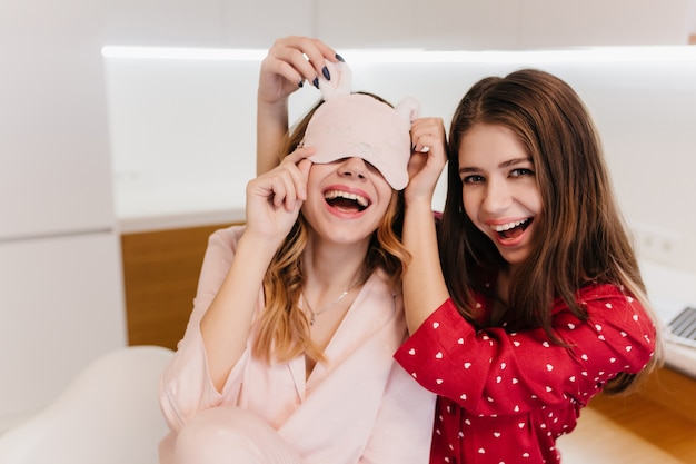 Romantic brown-haired girl wears red pajamas with smile. Photo of playful sisters in night-suits having fun.