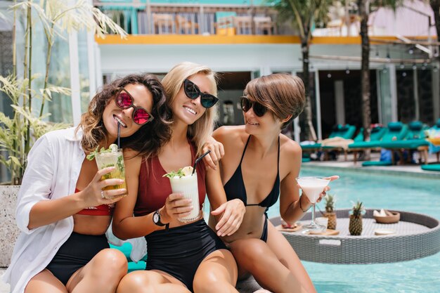 Romantic brunette woman in pink glasses drinking cocktail during photoshoot with friends. Fascinating ladies spending weekend in pool.