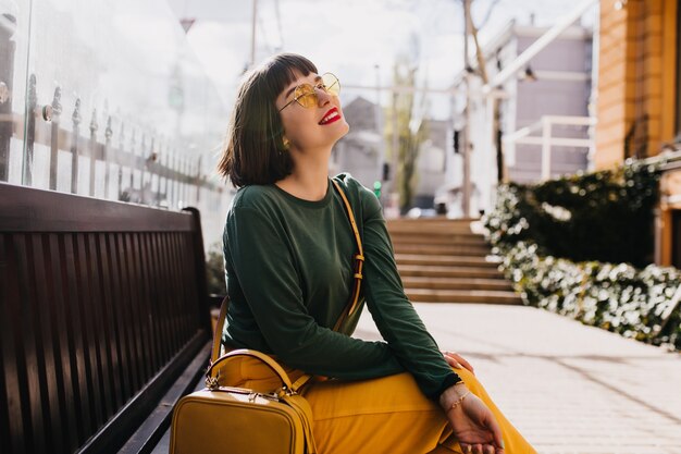 Romantic caucasian girl in trendy green sweater posing on street. Outdoor shot of blissful brunette female model chilling on bench.