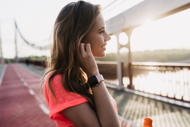 로맨틱 백인 여자는 경기장에서 포즈를 취하는 smartwatch를 착용합니다. 강 근처에서 아침을 보내는 즐거운 젊은 여자의 야외 샷.