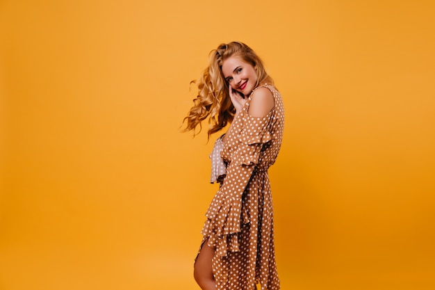 Romantic caucasian woman in brown dress with smile. Indoor photo of enchanting female model with blonde hair isolated on yellow wall.